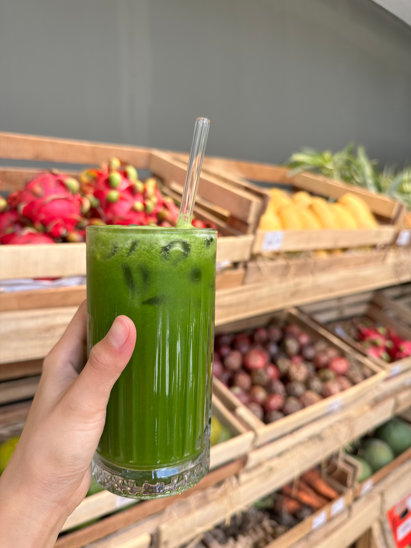 A green iced juice in front of fresh fruit shelves inside Tropi Farm